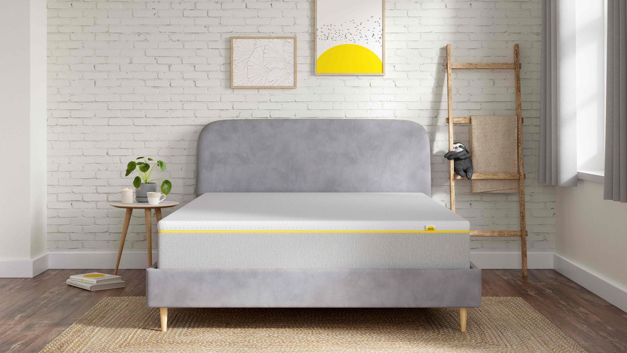 The eveSleep Wunderflip Lighter Hybrid mattress on a grey upholstered bed in a bedroom with a white painted brick wall