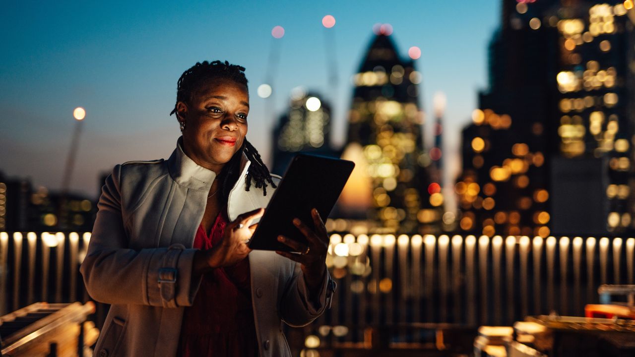 A businesswoman stands on the roof of a skyrise at sunset and looks at her tablet.