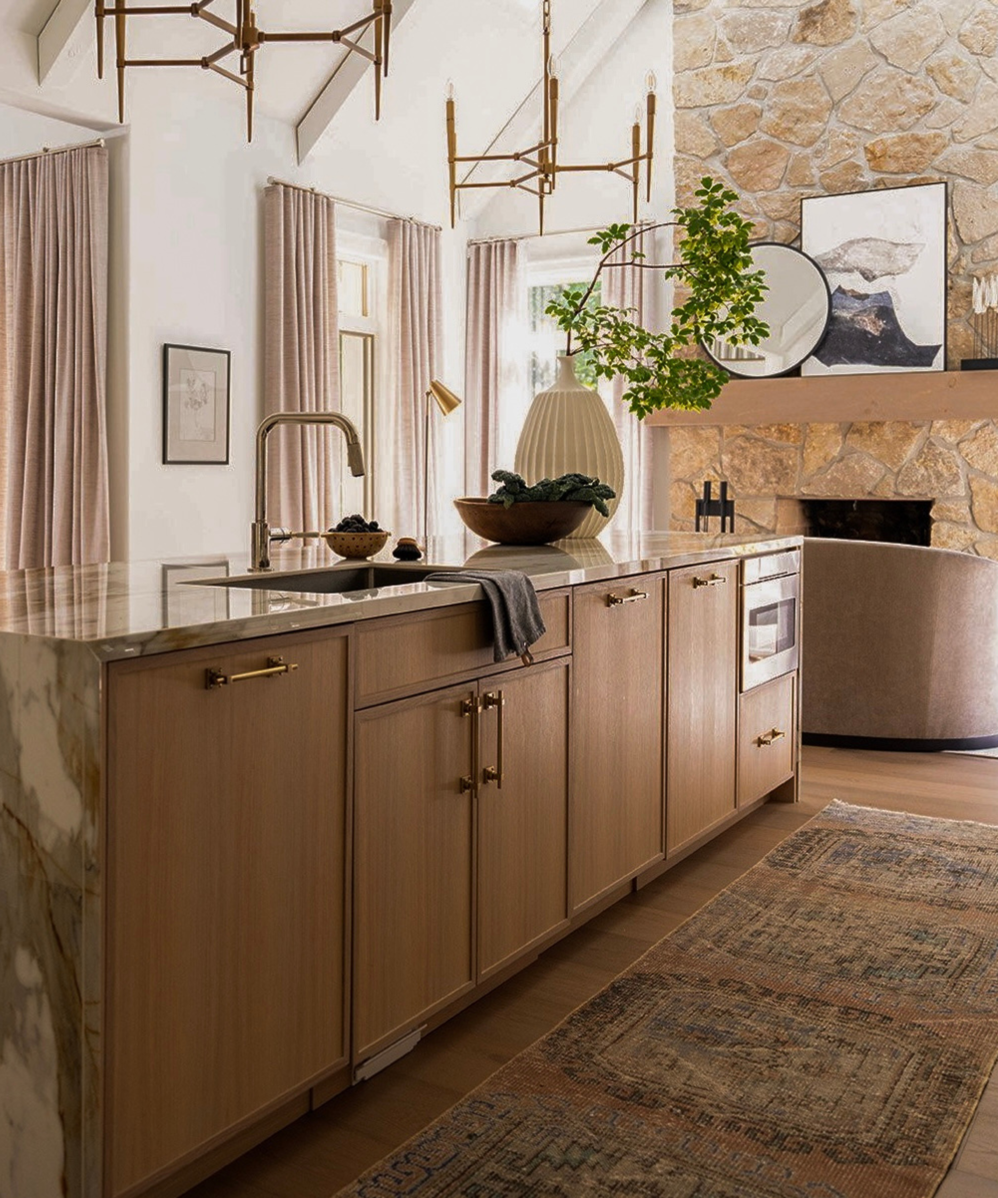 a kitchen island with light wood cabinets and a marble top surface which waterfalls over the side, a persian runner rug on the floor, and a large stone fireplace in the background