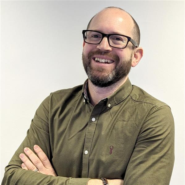 male with short dark hair and beard, wearing glasses and green shirt, stood with arms folded