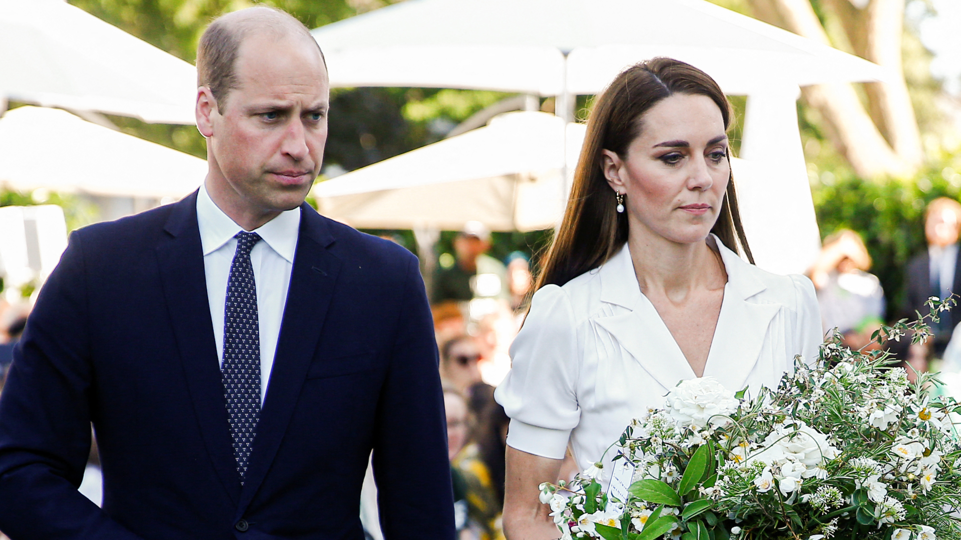 Prince William wears a suit and looks stern while Kate Middleton wears a white dress and carries a large bouquet of white flowers
