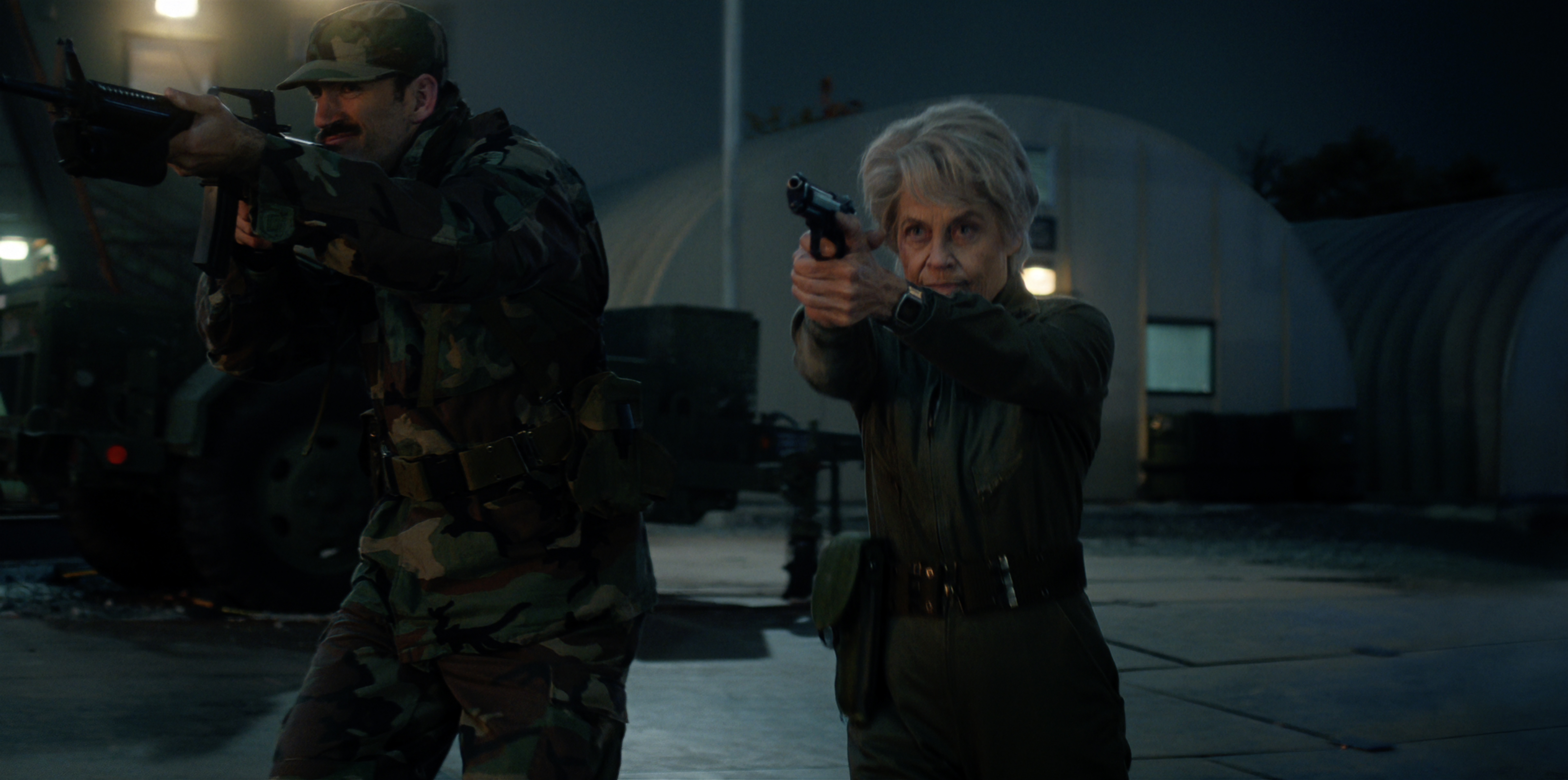 STRANGER THINGS SEASON 5 still of Linda Hamilton as Dr. Kay wearing all black pointing a gun on the grounds of a military facility as she walks with another soldier
