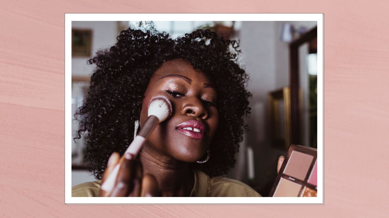 A woman pictured applying powder blush to the apples of her cheeks with a blusher brush/ in a pink template