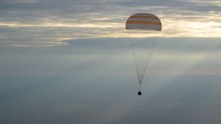 a space capsule descended through the dawn sky under a parachute, approaching a landing in the desert