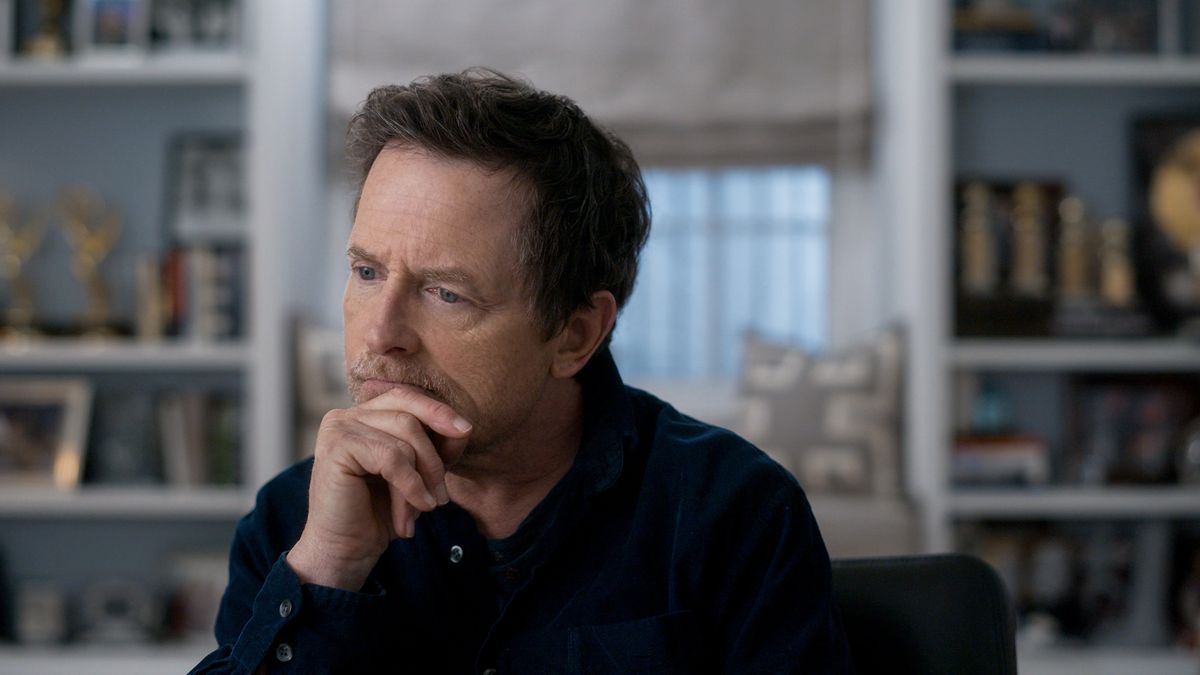 A scene from Still: A Michael J Fox Movie where Michael J Fox is being interviewed, facing into the camera. He is resting his chin on his right hand and looking off to the side slightly pensively