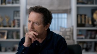 A scene from Still: A Michael J Fox Movie where Michael J Fox is being interviewed, facing into the camera. He is resting his chin on his right hand and looking off to the side slightly pensively