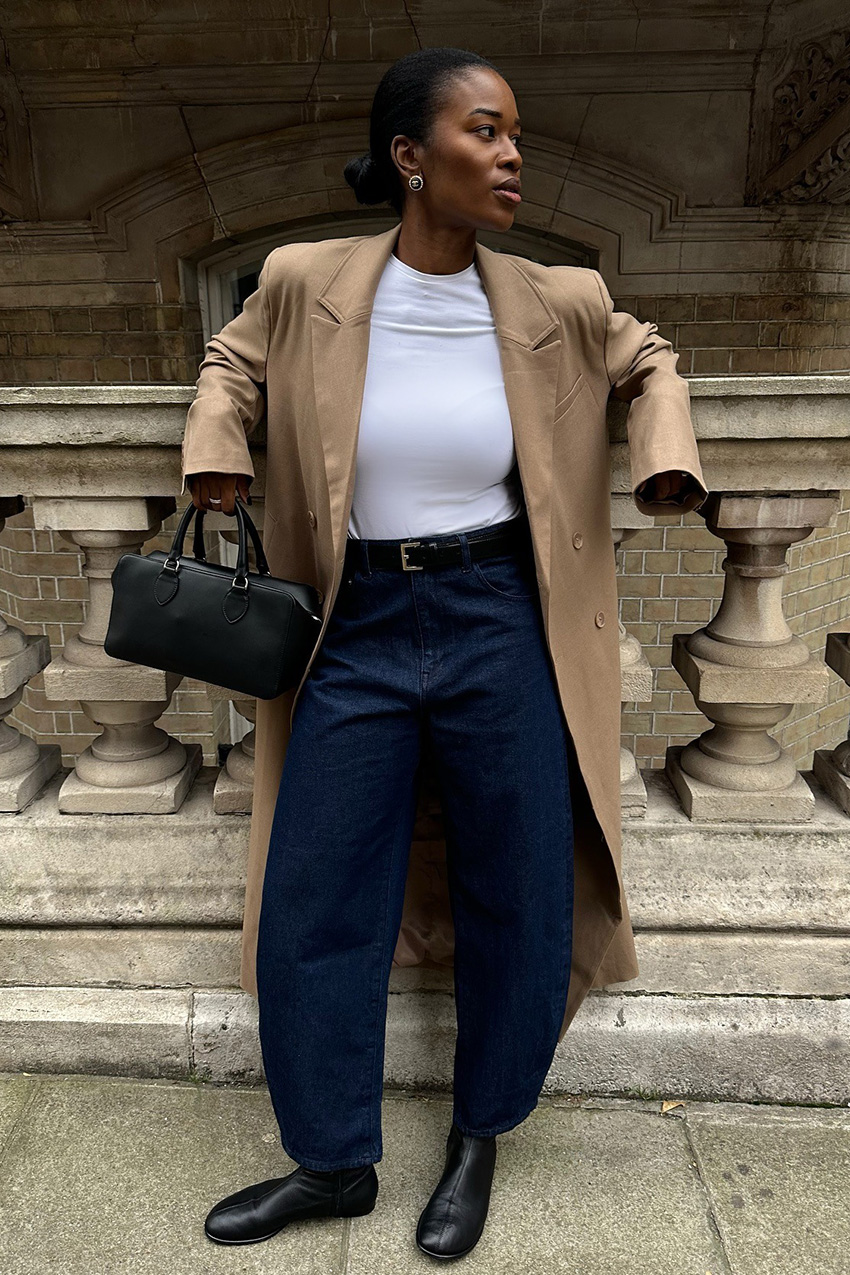 Denim and shoe combos shown in a street style photo of a woman standing outside wearing a long tan coat over a white t-shirt styled with dark wash barrel leg jeans, black flat sock boots, a black belt, silver earrings, and a black bowler bag
