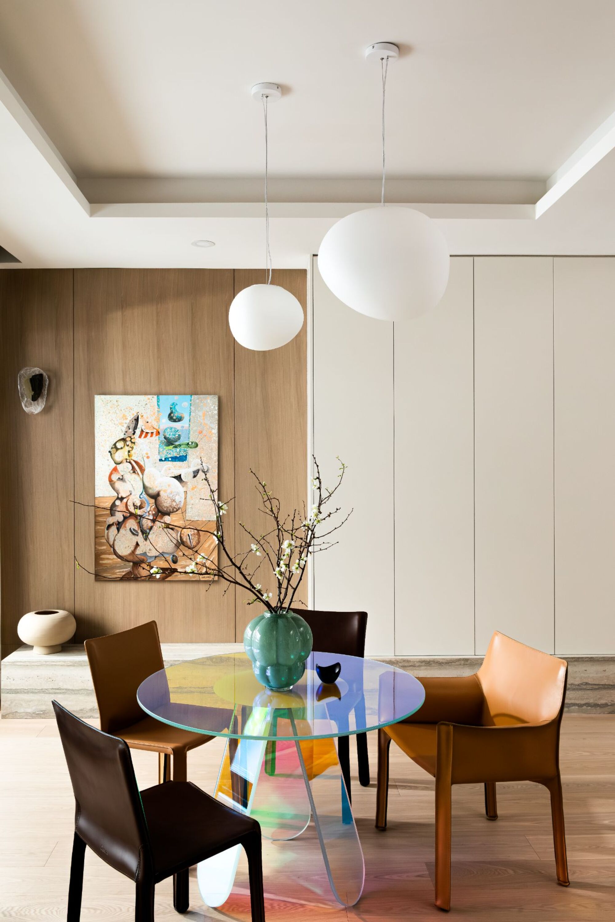 A dining area with an iridescent, round table and brown leather chairs around. Behind is a large surrealist artwork on the wooden clad walls.