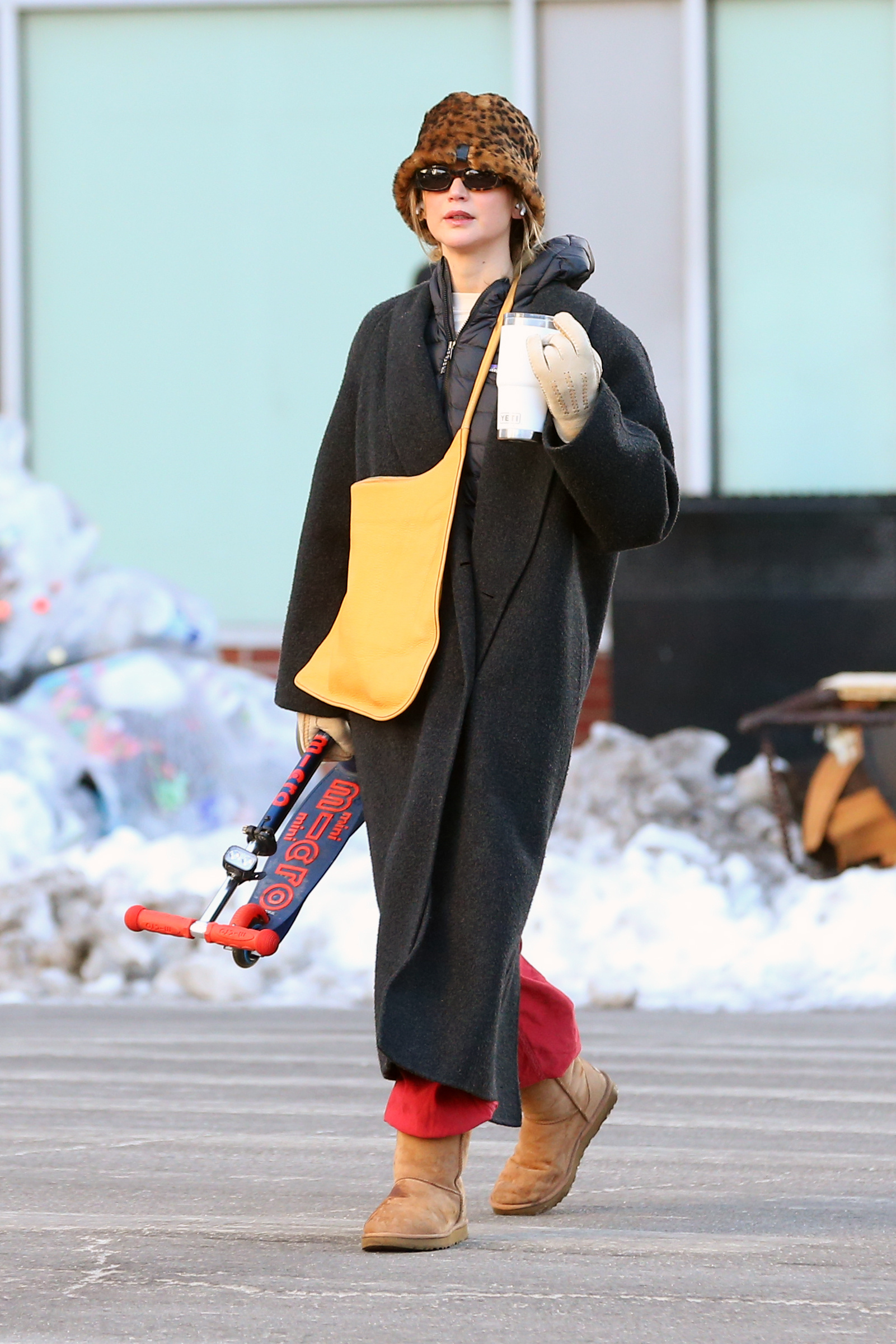 Jennifer Lawrence walks down the street wearing a black coat with Uggs, red trousers and a leopard print hat.