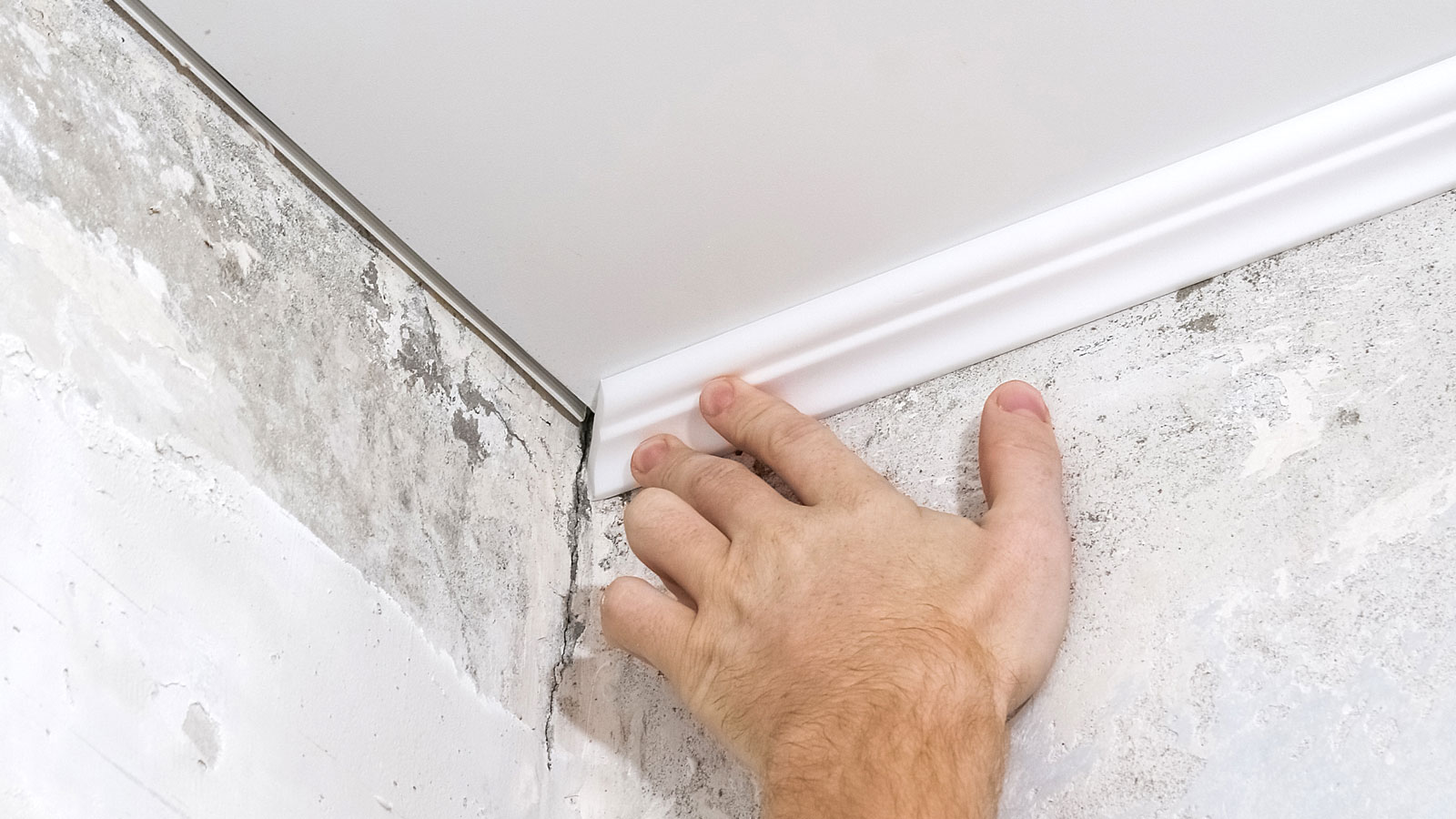 Hand holding narrow white coving against corner of bare plaster wall and ceiling