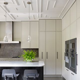 off white and grey kitchen with white panelled ceiling, kitchen island with marble top, dark grey bar stools, grey mosaic splash back, glass pendants, fern on island