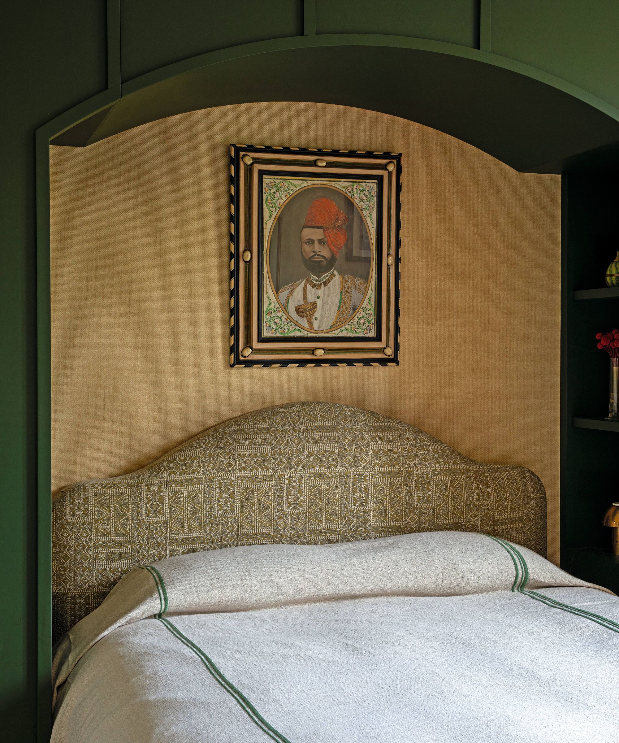 a green painted arched bed alcove nook dressed with grasscloth paper, a patterned curved headboard, white and green linens and artwork hung above