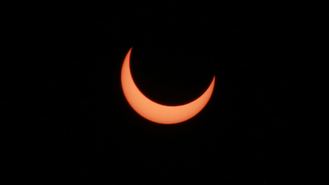 An upward-facing crescent of the sun's disk is visible behind the silhouette of the moon against a black sky during a solar eclipse.