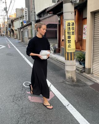 Clara wears a black sweater, black midi skirt, and black flip flops.