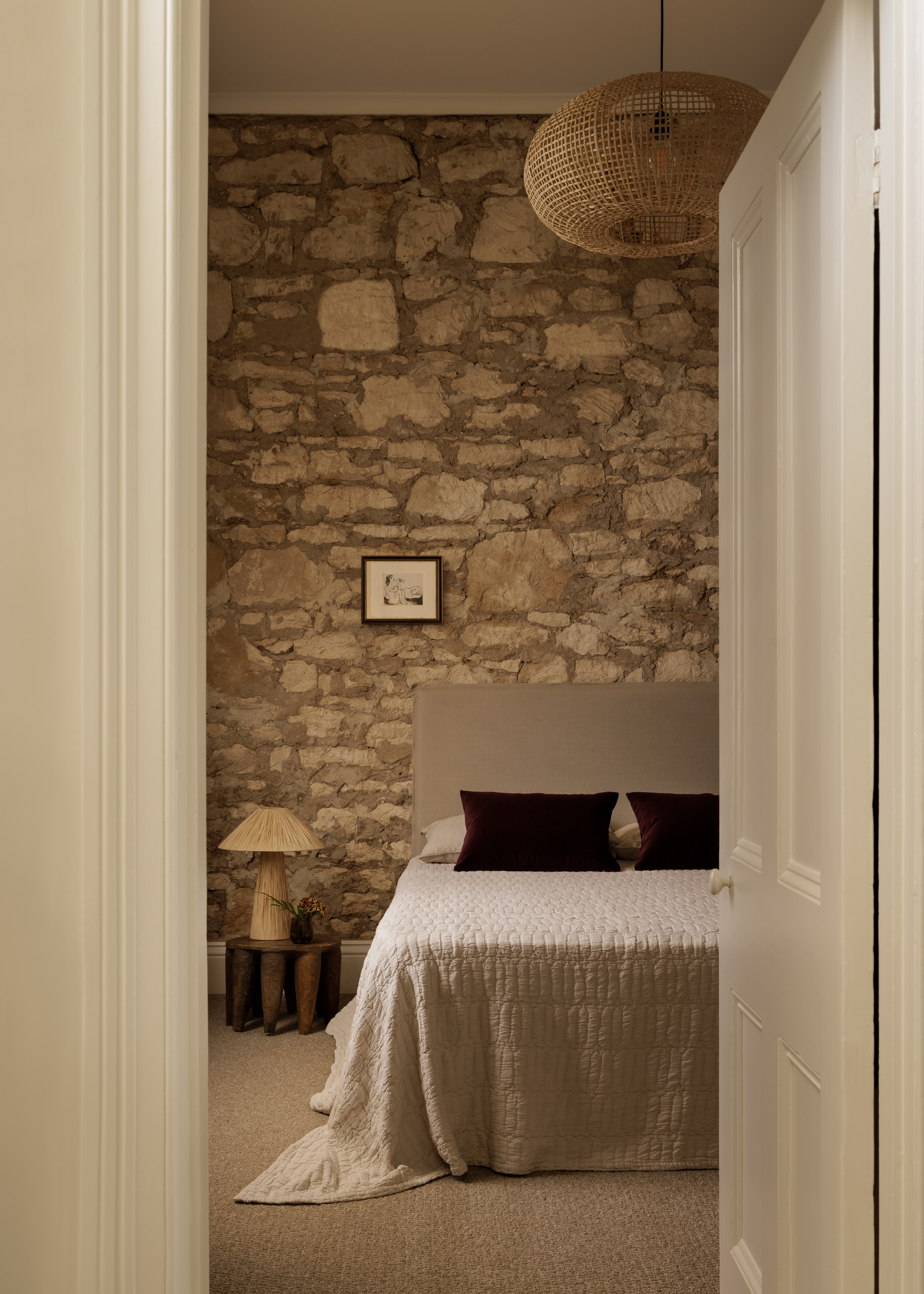 A stone bedroom wall with a hanging woven pendant lamp, a low gray bed with a pair of purple pillows, beside a low wood stool with a woven lamp and a vase