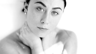 A close-up of a person cupping their neck, wearing earrings and multiple rings, set against a soft, blurred white background