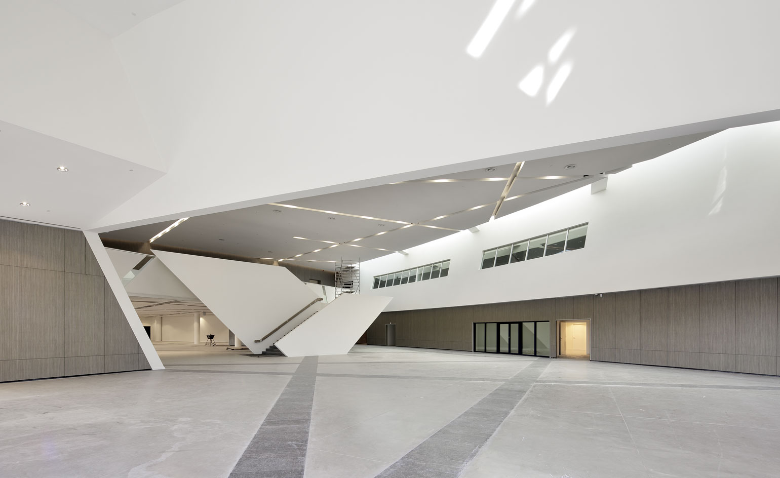Inside the centres large foyet, grey marble floor design, three connected black framed sliding doors on the right hand wall, with windows sectioned above on the first floor level, white ceiling, grey walls and ceiling lights in the middle section of the large space