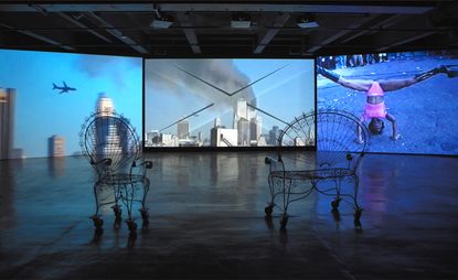Two wire frame chairs facing three wall size tv screens on a concrete floor.