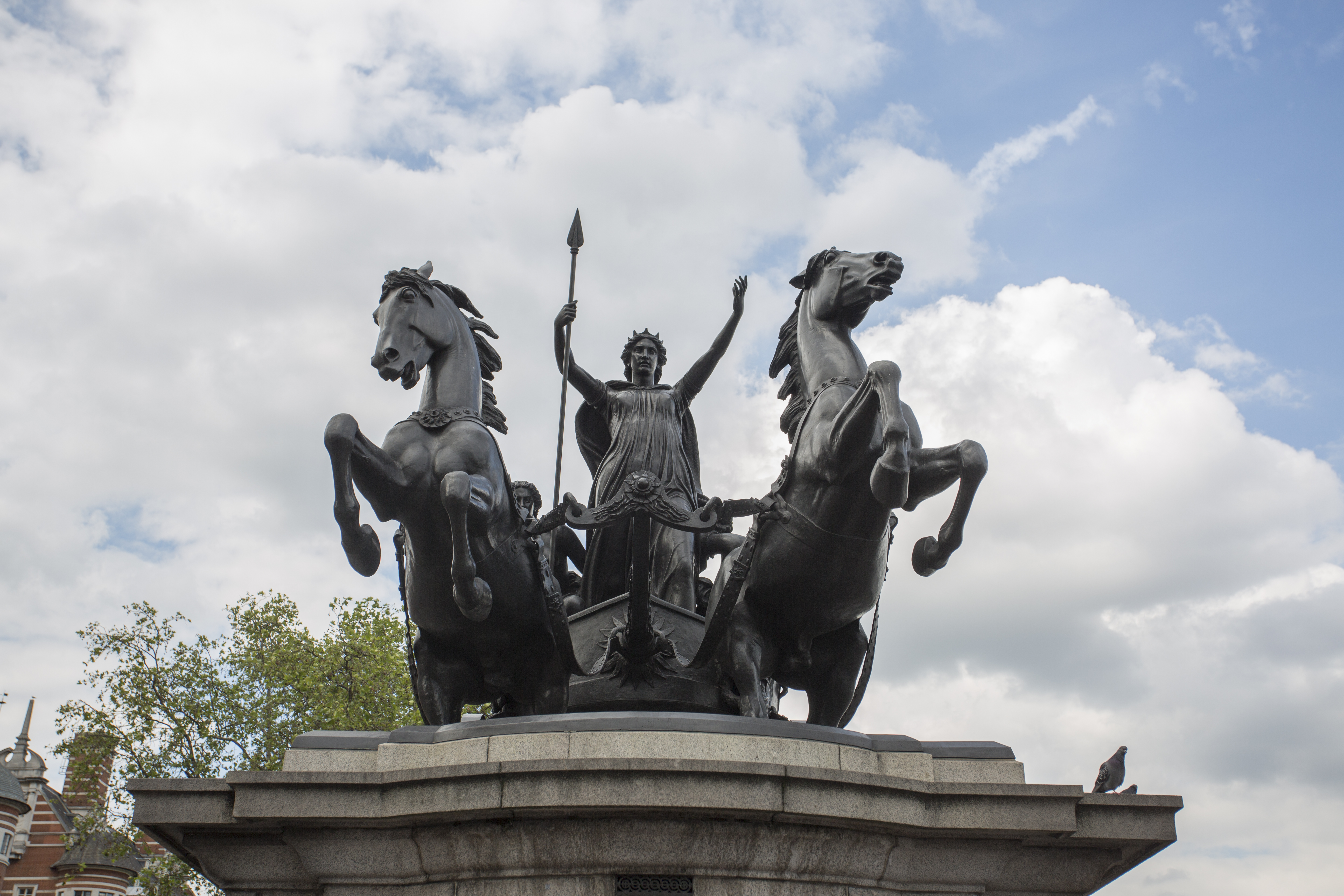 Bronze statue of Boudica on a chariot led by horses, shot from below with cloudy sky in the background