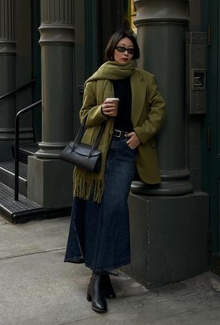 A denim skirt outfit is shown in a photo of a woman on the sidewalk wearing an oversize green blazer with a matching scarf, a black sweater, a black belt, a denim mini skirt, black boots, and a black shoulder bag