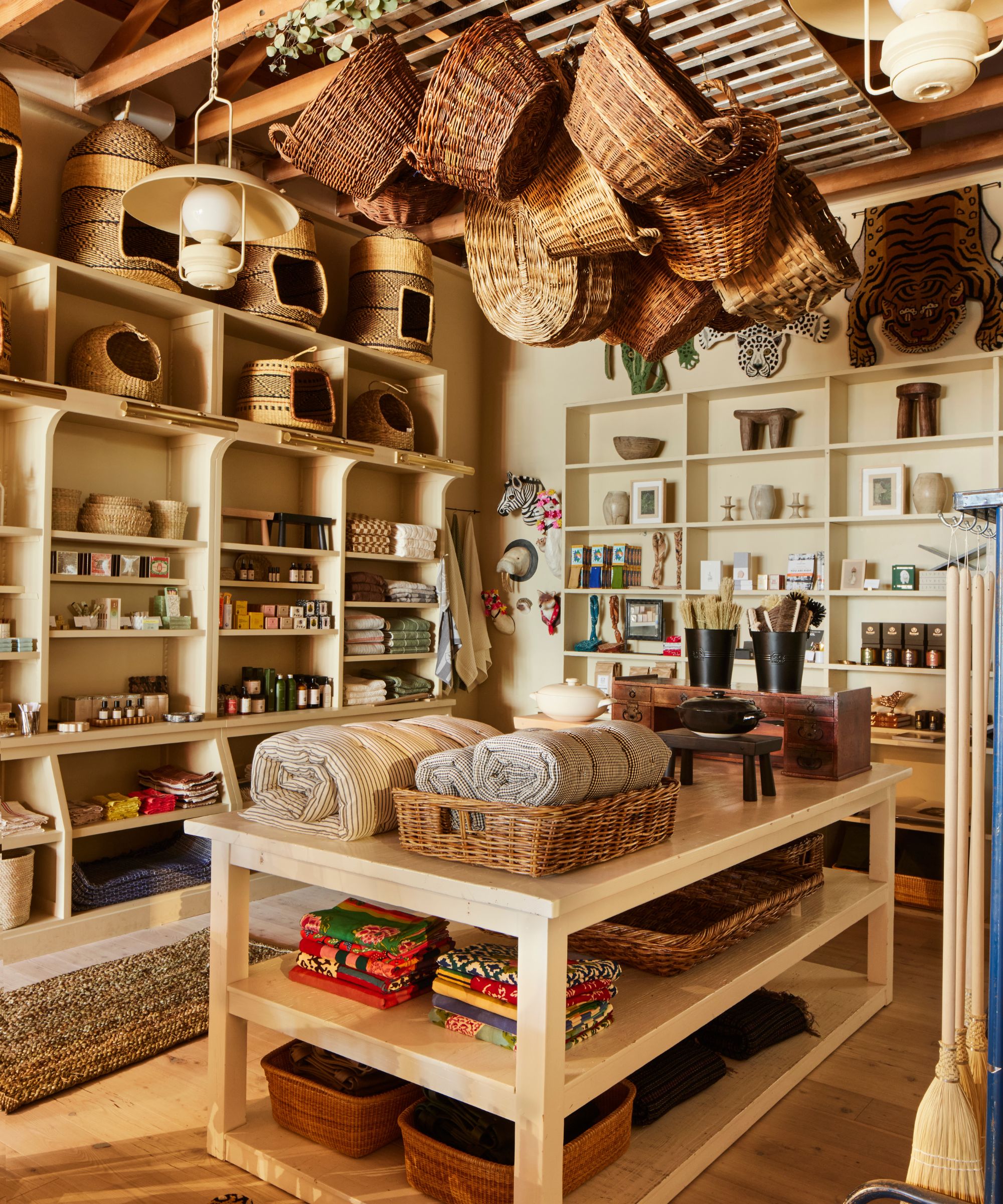 Inside a homeware store with open shelving painted cream, wood island with merchandise and baskets hanging on ceiling