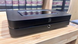 Bluesound Node (2024) music streamer slight angled view on wooden hi-fi rack in front of books