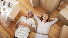 A smiling older couple relax on the floor, surrounded by moving boxes.