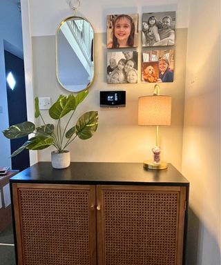 A sideboard in black and wicker with a cream and gold lamp, showing a mirror and four familiy photos on the wall, nestled around a black Vaillant digital thermostat