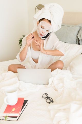 A woman sits on her bed in front of her laptop wearing a face mask while painting her nails