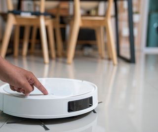 A finger switching on a robot vacuum to clean a kitchen floor.