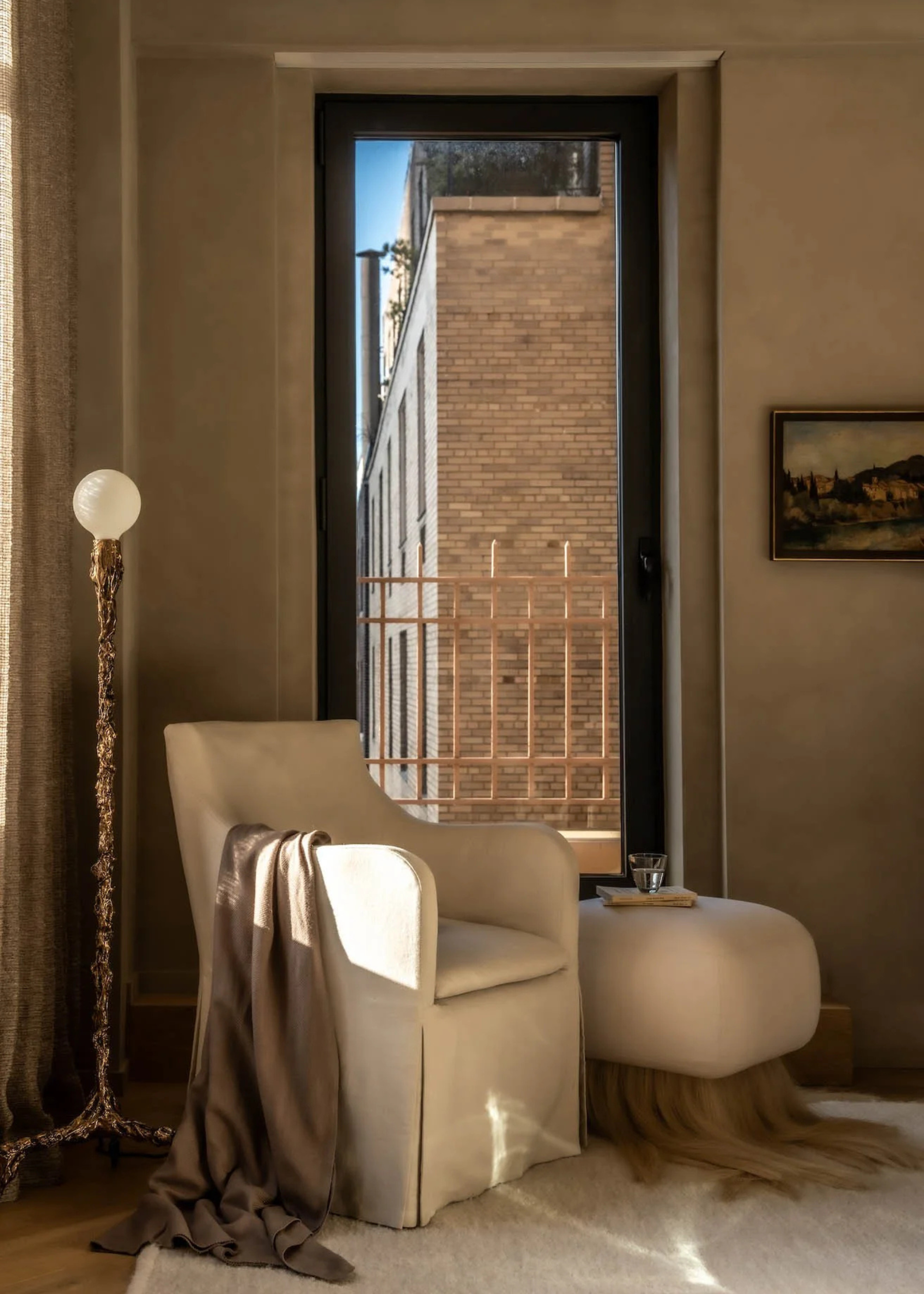 A sandy beige living room corner with a gilded floor lamp near a minimalist sofa seater and a pouf with a faux silky fringe