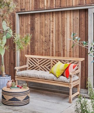 wooden garden bench with padded seat cushion on a patio in front of a wood clad wall