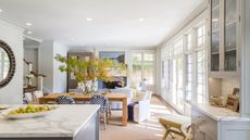 Open plan living room and kitchen with crittall doors, cabinetry painted in Benjamin Moore's Gray Owl, a marble island, a dining table with a vase of foliage, and a white sofa and armchair.