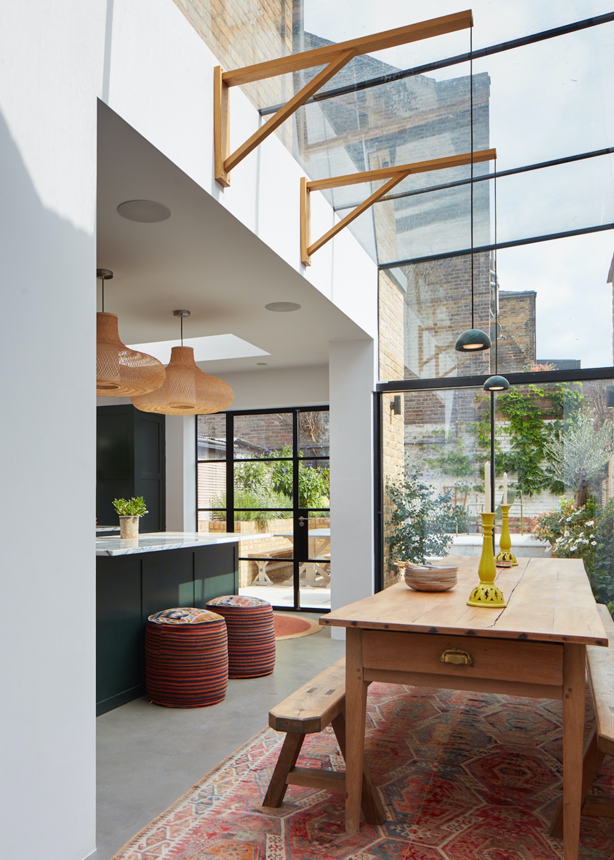 A glass kitchen extension with an island and a wooden oak dining table. On the dining table are two candles and beside the table are two bench stools. The kitchen also has large windows/doors looking into the backyard.