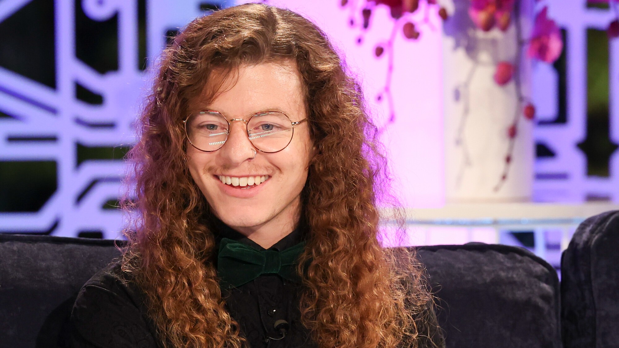Quinn Martin smiling during the jury roundtable segment on Big Brother. There is a purple and black background behind him and a pink plant.