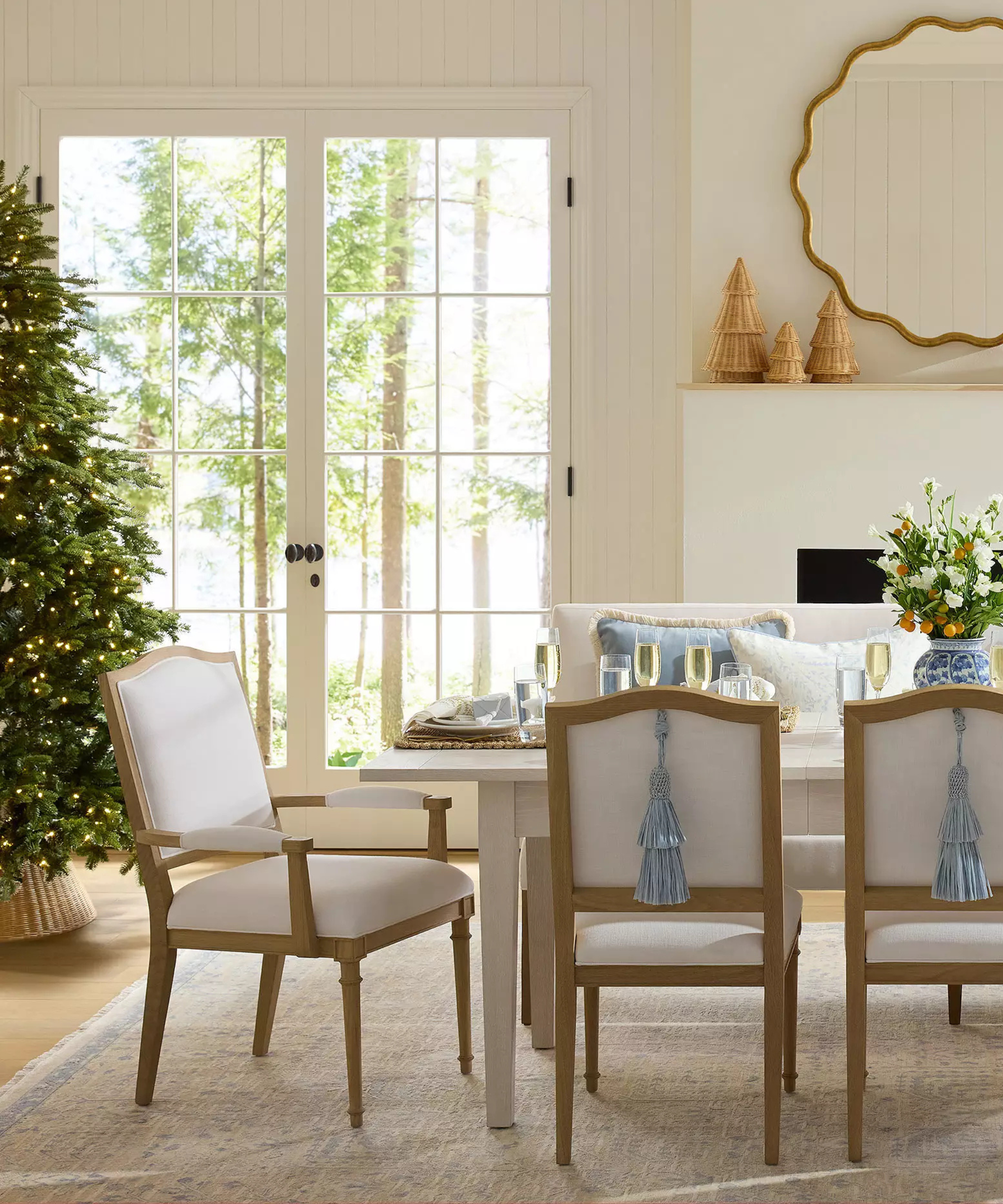 a festive dining room with a tree to the left, a table with chairs in the middle, and a mantel with a mirror above and a collection of three rattan trees on one side