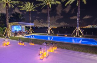 A night view of a pool area with, a grass lawn, palm trees, floor lamps and a sea view.