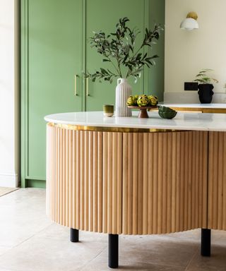 A kitchen island with a wooden panel base, a white surface with green plants and dinnerware on, and a light green cabinet behind it