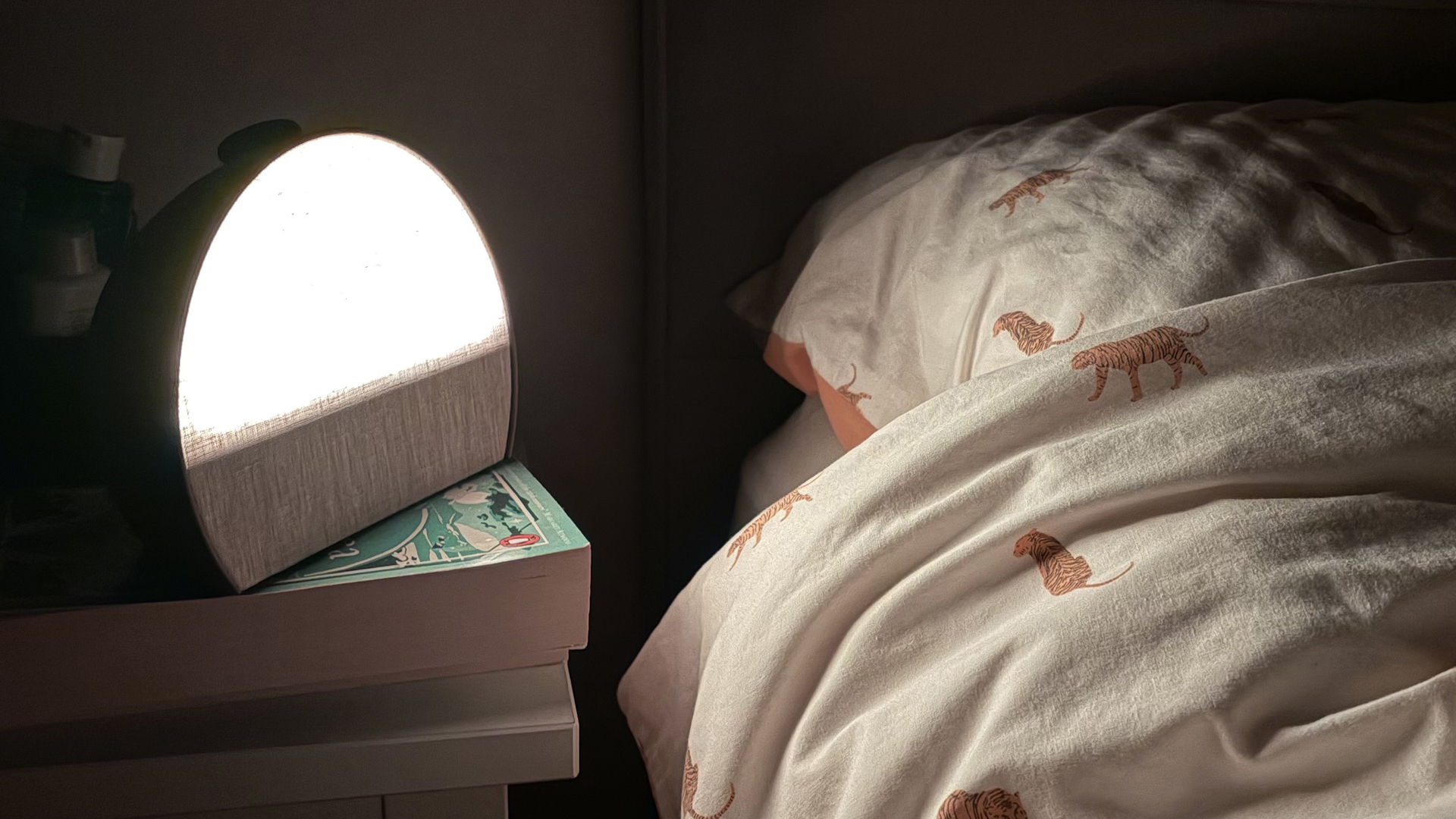 The Hatch Sleep Clock on a nightstand next to a bed in the reviewer's bedroom. The Hatch is glowing which is lighting up the bed