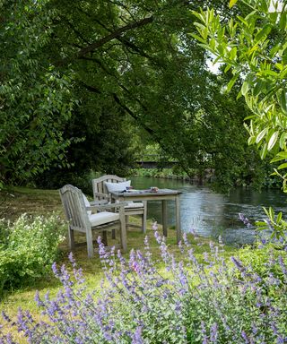 A wooden dining table with two chairs next to a river in front of lavender