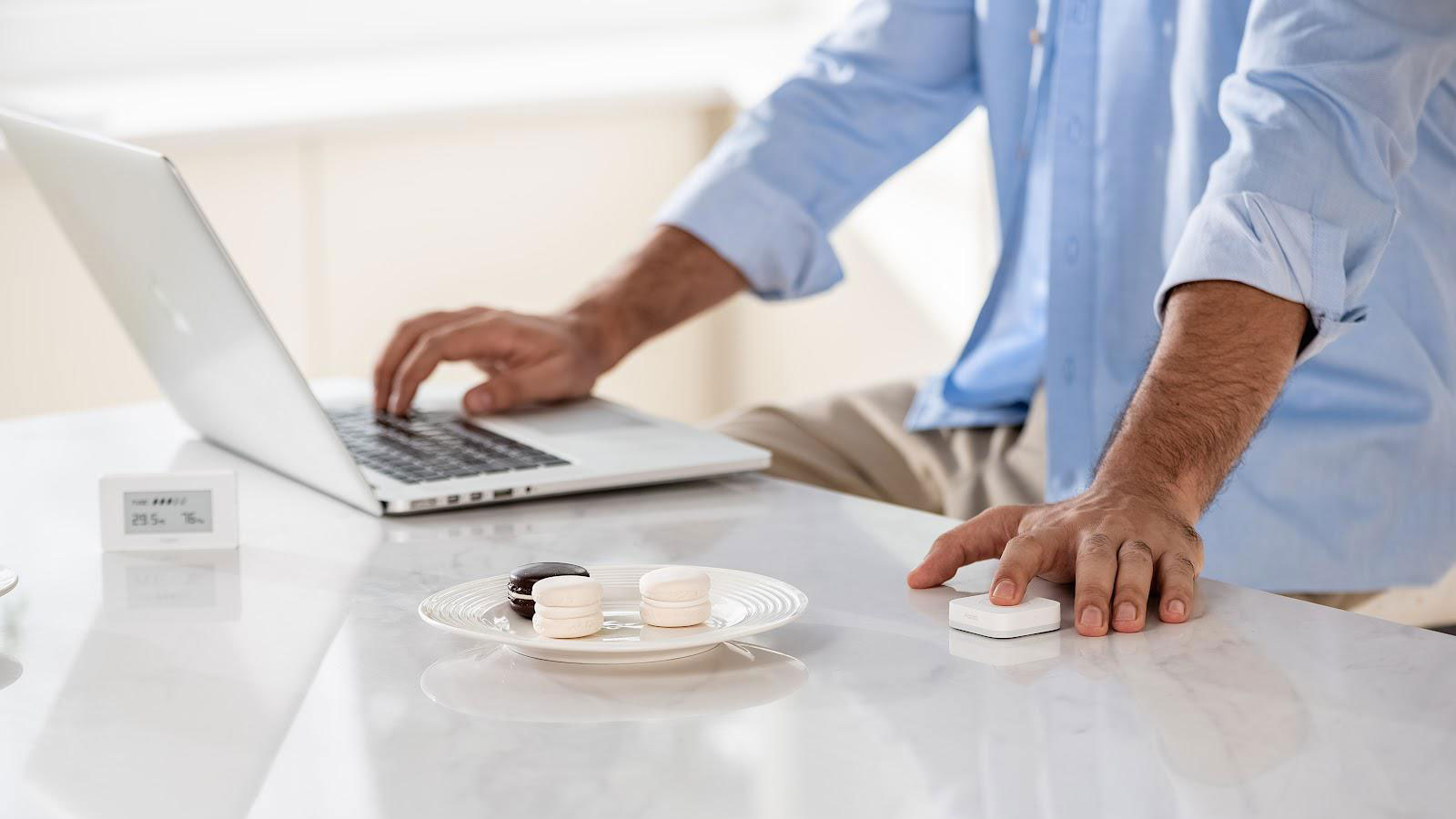 Los brazos de un hombre están en tiro.  Está trabajando en su computadora portátil con una mano y administrando su hogar inteligente con la otra.