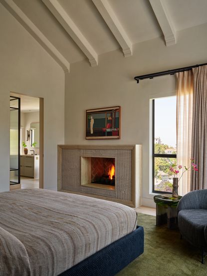 Image of a white bedroom with high, angled ceilings and a fireplace and large window on the back wall. There are layered window treatments on the window. There is a green rug under the bed, and a chair and side table in the corner.