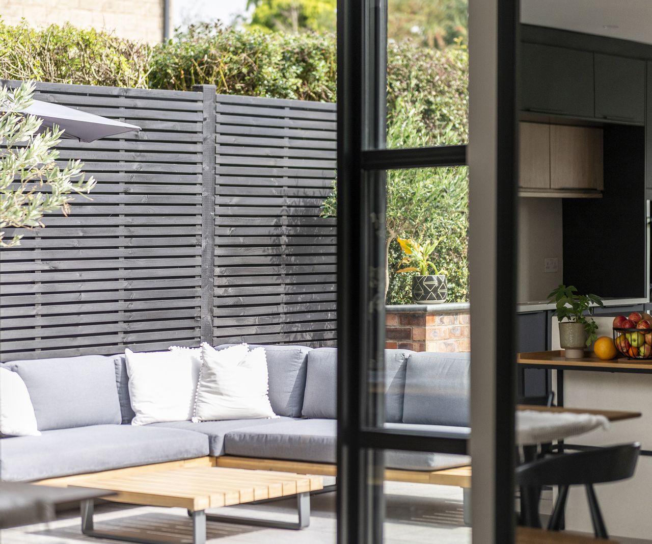 A view of a patio with grey outdoor corner sofa in front of a charcoal grey fence. The area is viewed through open glass patio doors
