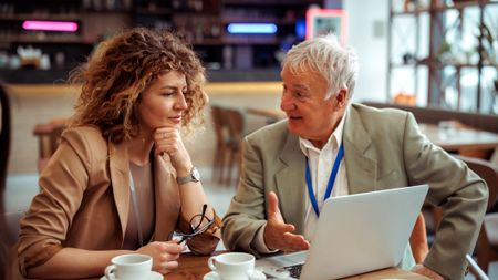 An older worker or consultant talks with a younger colleague at a cafe. They are both wearing lanyards, as if at a conference.