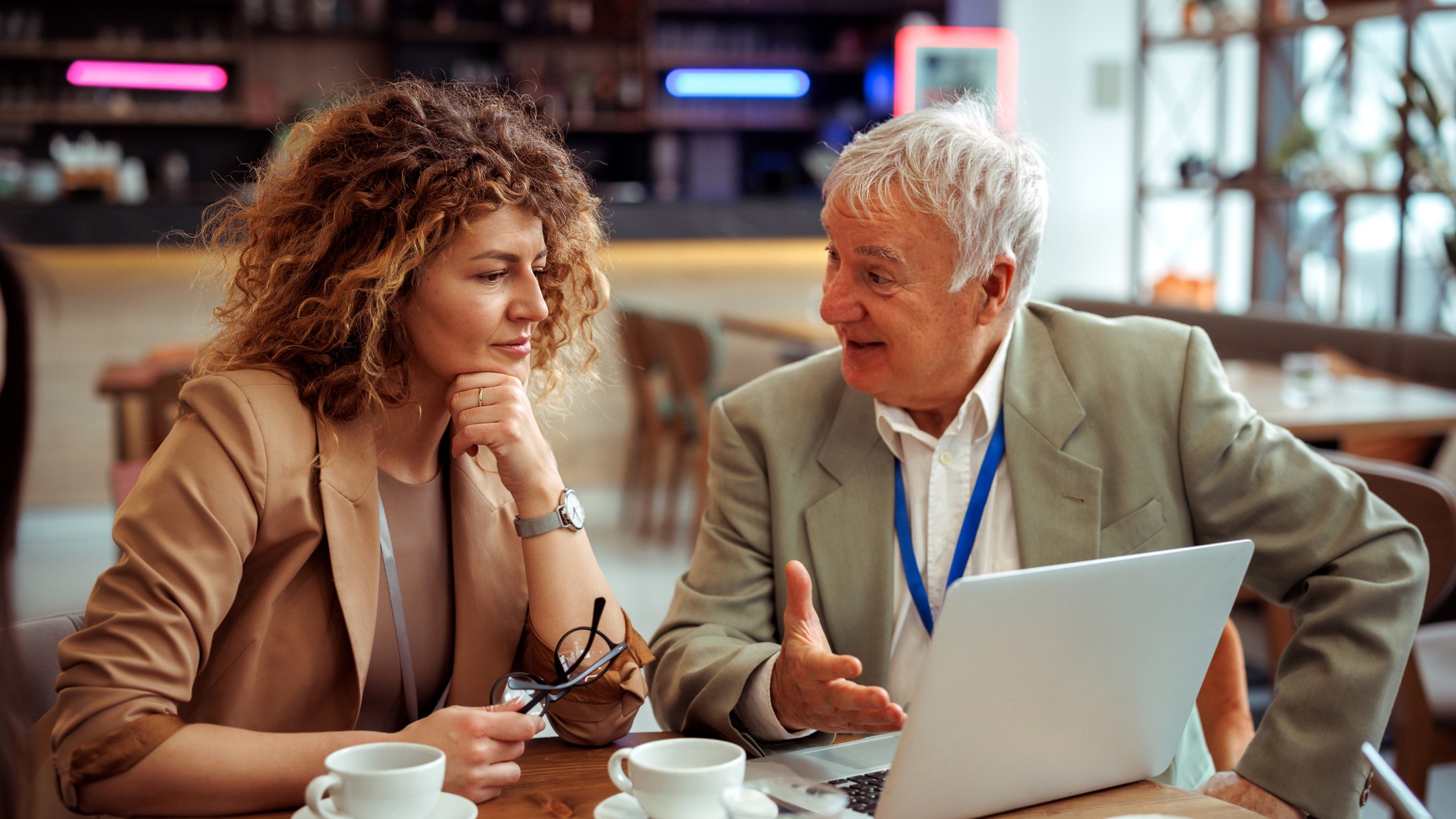 An older worker or consultant talks with a younger colleague at a cafe. They are both wearing lanyards, as if at a conference.
