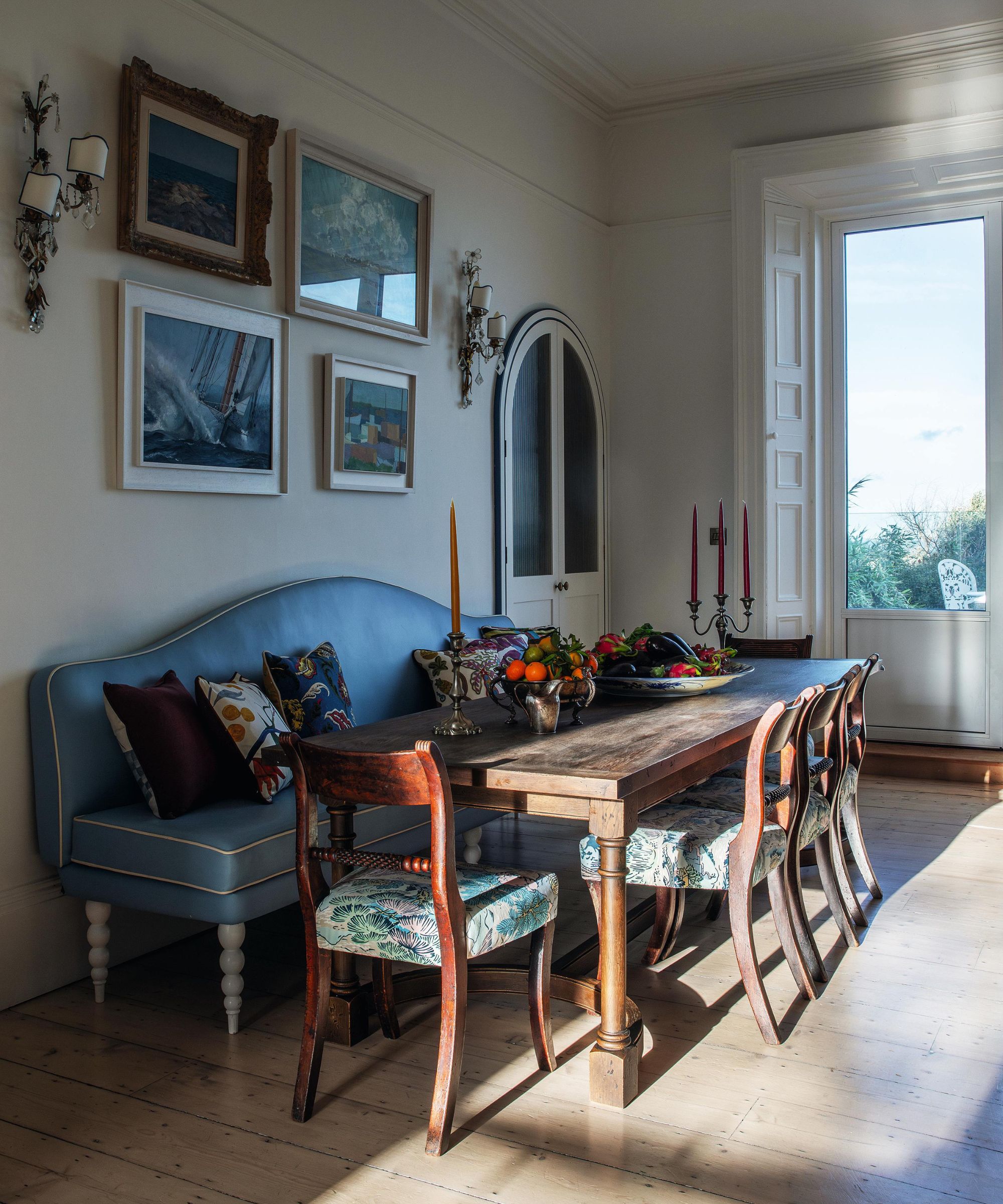 a white kitchen with large window out to the garden with a blue banquette seat and antique wooden table and chairs