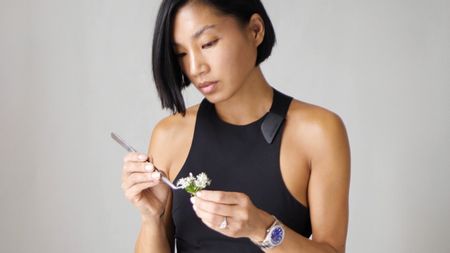 A young woman in a black tank top and silver jewelry, including a silver watch, uses a silver fork to perfect a floral arrangement while standing against a grey background.
