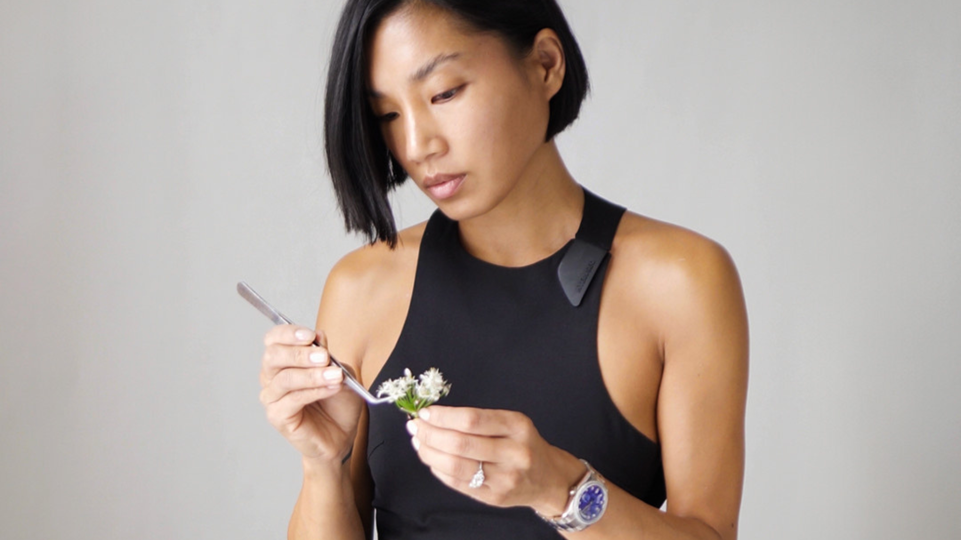 A young woman in a black tank top and silver jewelry, including a silver watch, uses a silver fork to perfect a floral arrangement while standing against a grey background.