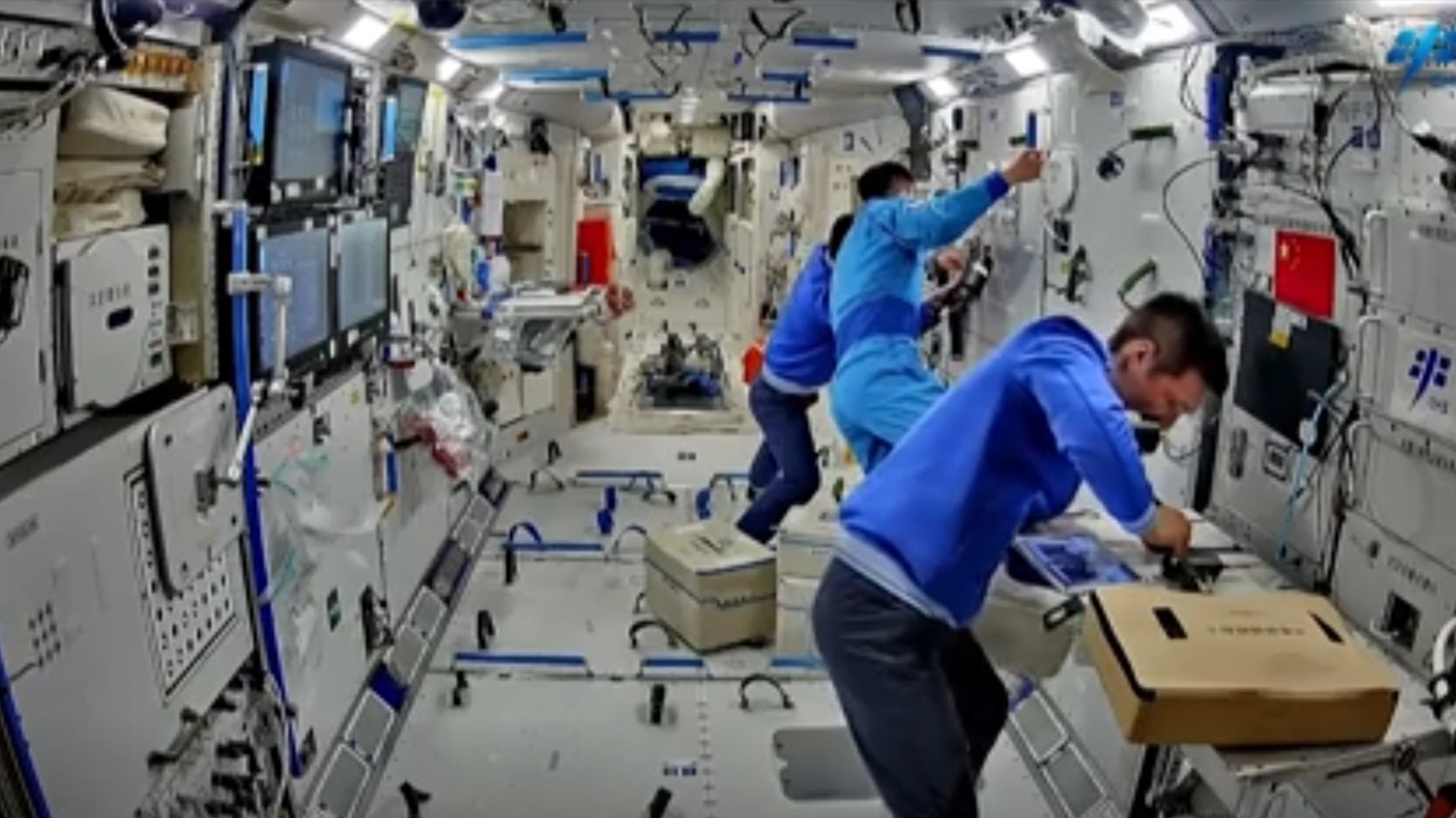 three astronauts in blue flight suits work inside a white-walled space station