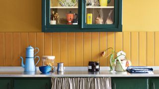 Bold kitchen with mustard yellow walls and deep green cabinetry. A glass-fronted wall cupboard displays colourful crockery and a yellow teapot.